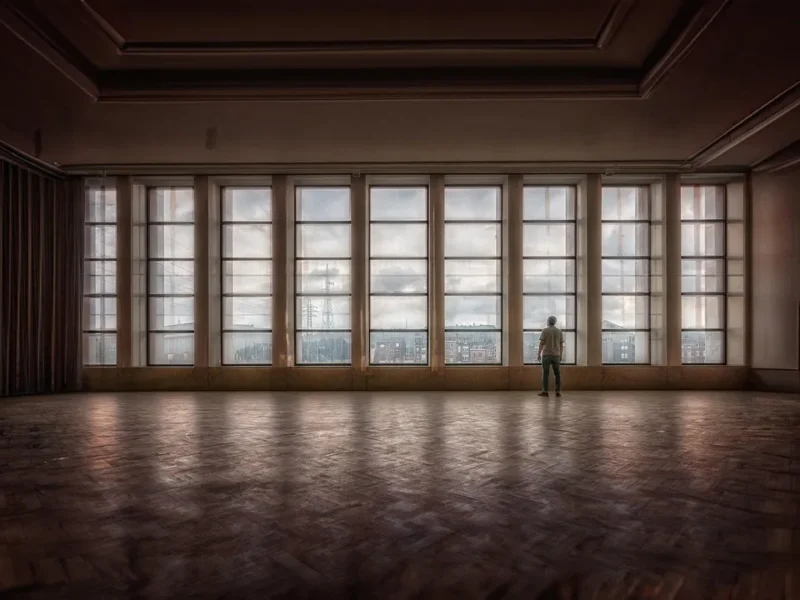 Pusta sala balowa z dużymi oknami i samotną postacią patrzącą przez nie