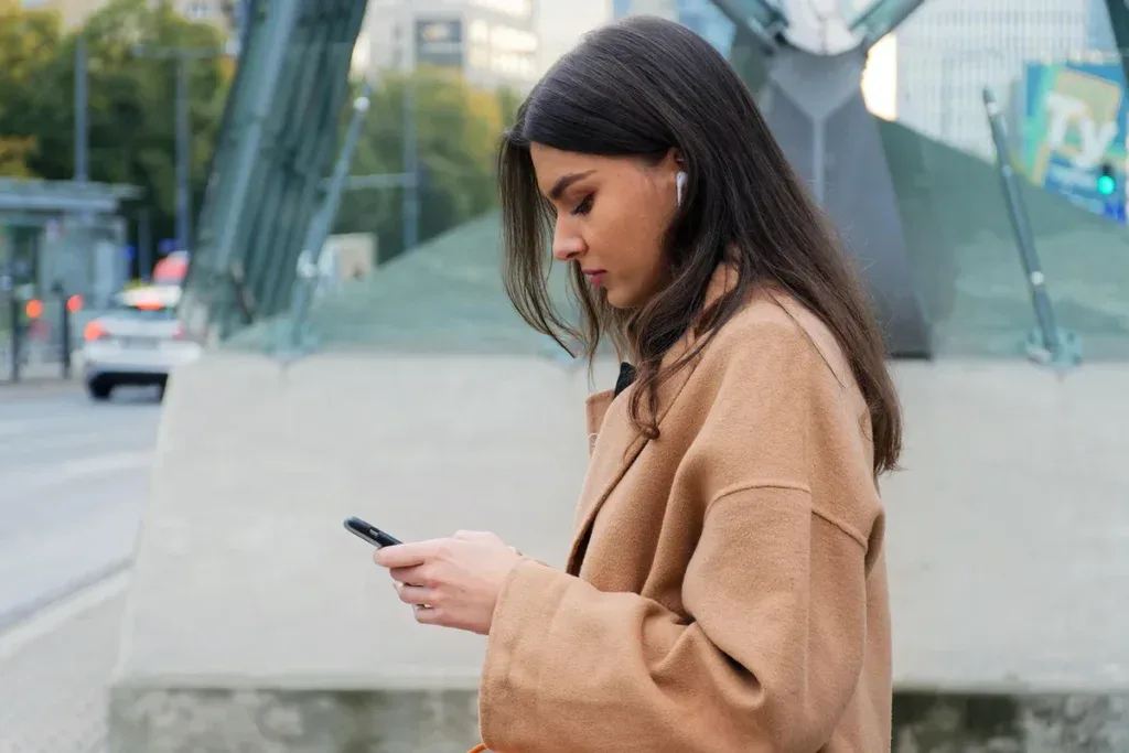 Kobieta w płaszczu wpatruje się w telefon na ulicy i czeka na wiadomość od partnera
