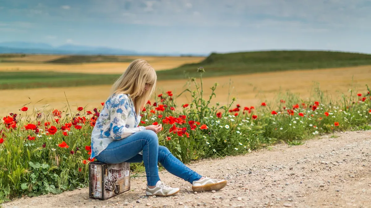 Trauma bonding: kobieta siedzi na walizce przy polnej drodze i waha się, czy odejść